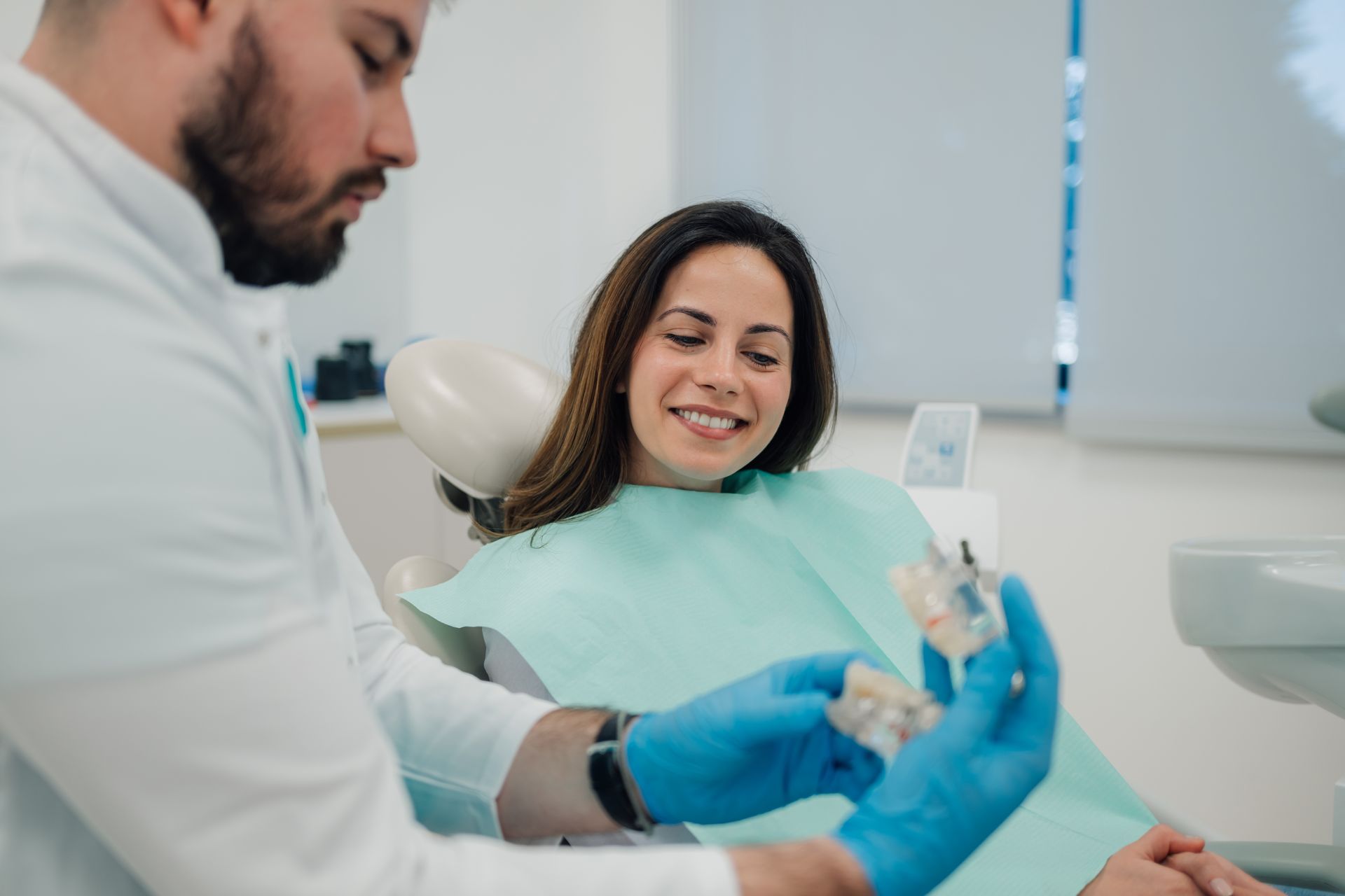 Dentist examining a smiling patient in a bright clinic while holding a dental tool.