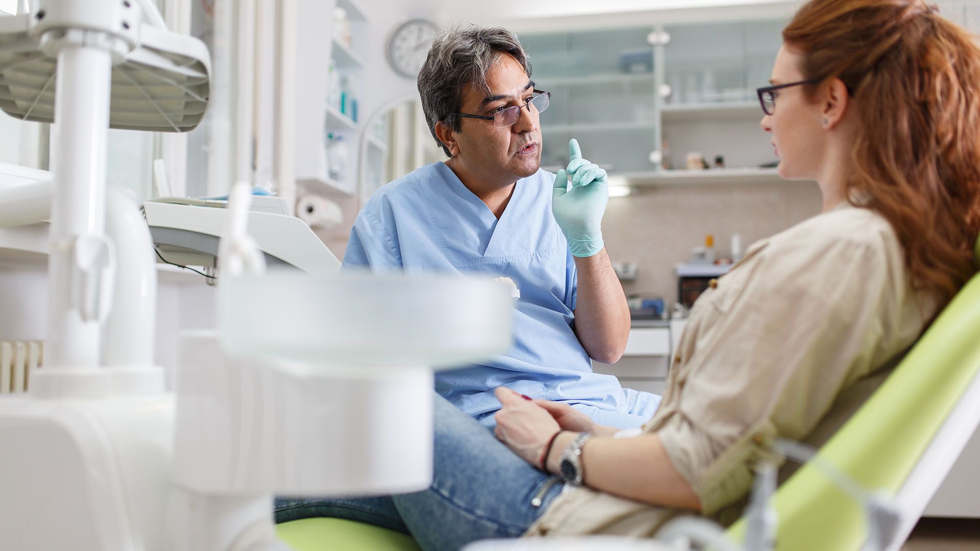Dentist in blue scrubs pointing while speaking to a female patient in a dental chair; clinic setting.