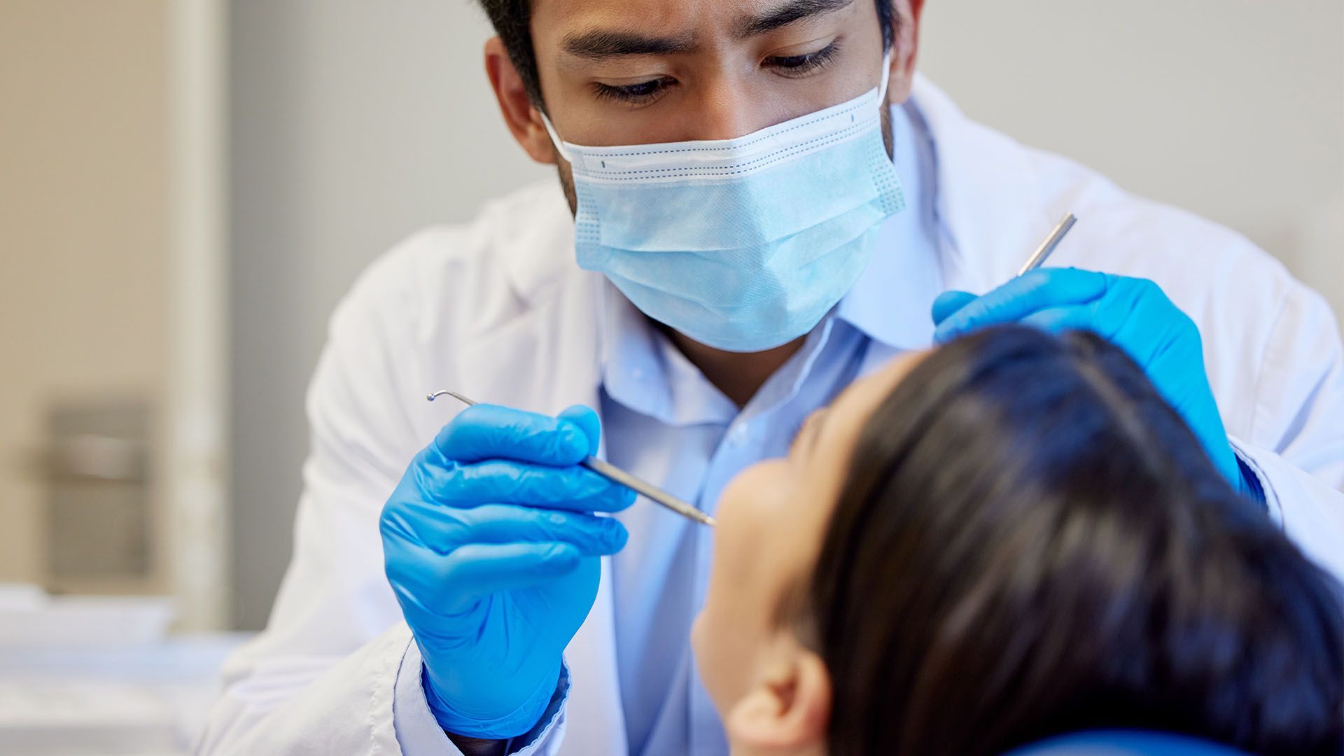 Dentist examining a patient's teeth. The dentist wears a mask, gloves, and a lab coat. The patient sits in a dental chair.