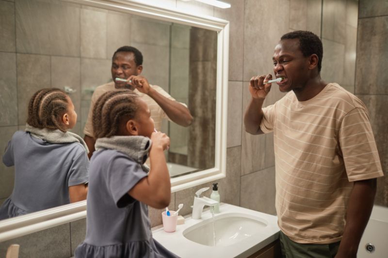 A man and a little girl are brushing their teeth in front of a bathroom mirror.