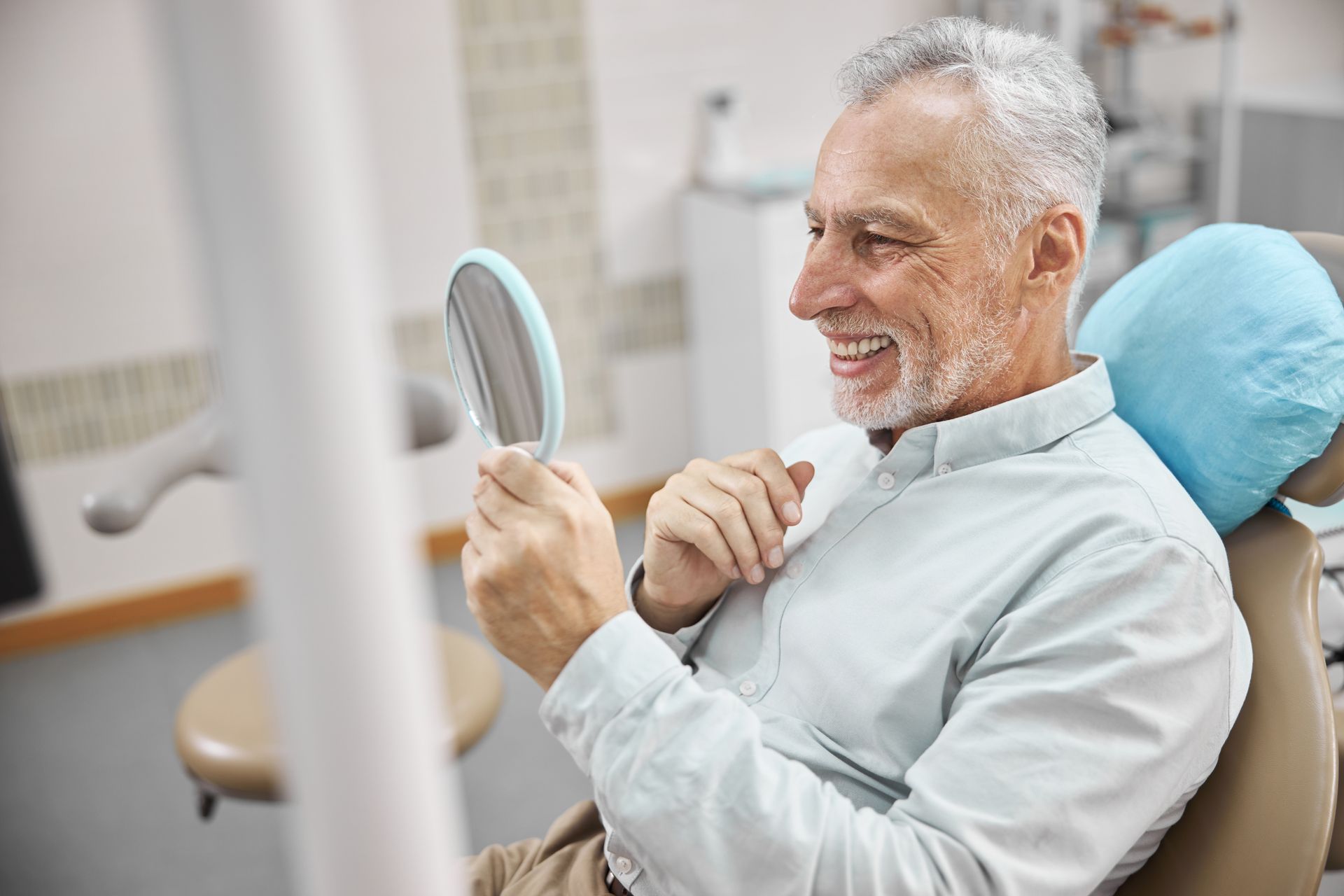 Man in dental chair smiles while looking at teeth in mirror.