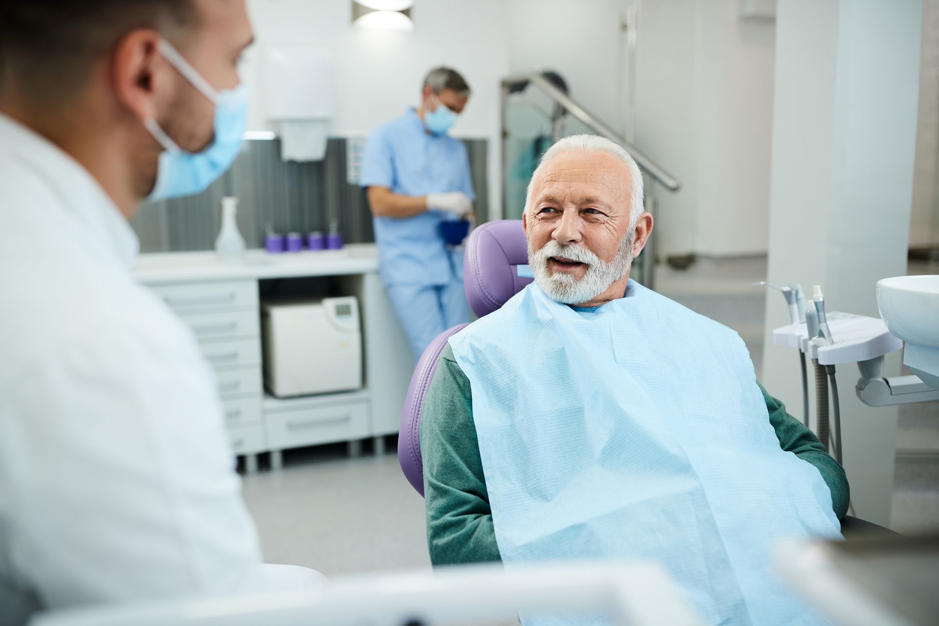 Dentist and patient in a dental clinic. The older patient is talking while the dentist is listening. A nurse is in the background.