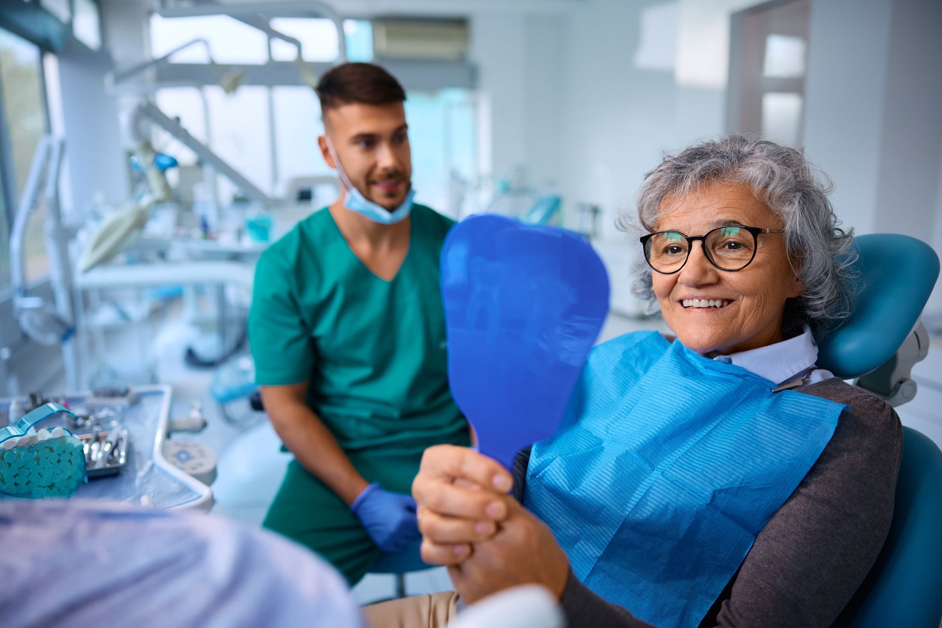 Smiling patient holding a blue mirror in a dental clinic, with a dentist in green scrubs nearby
