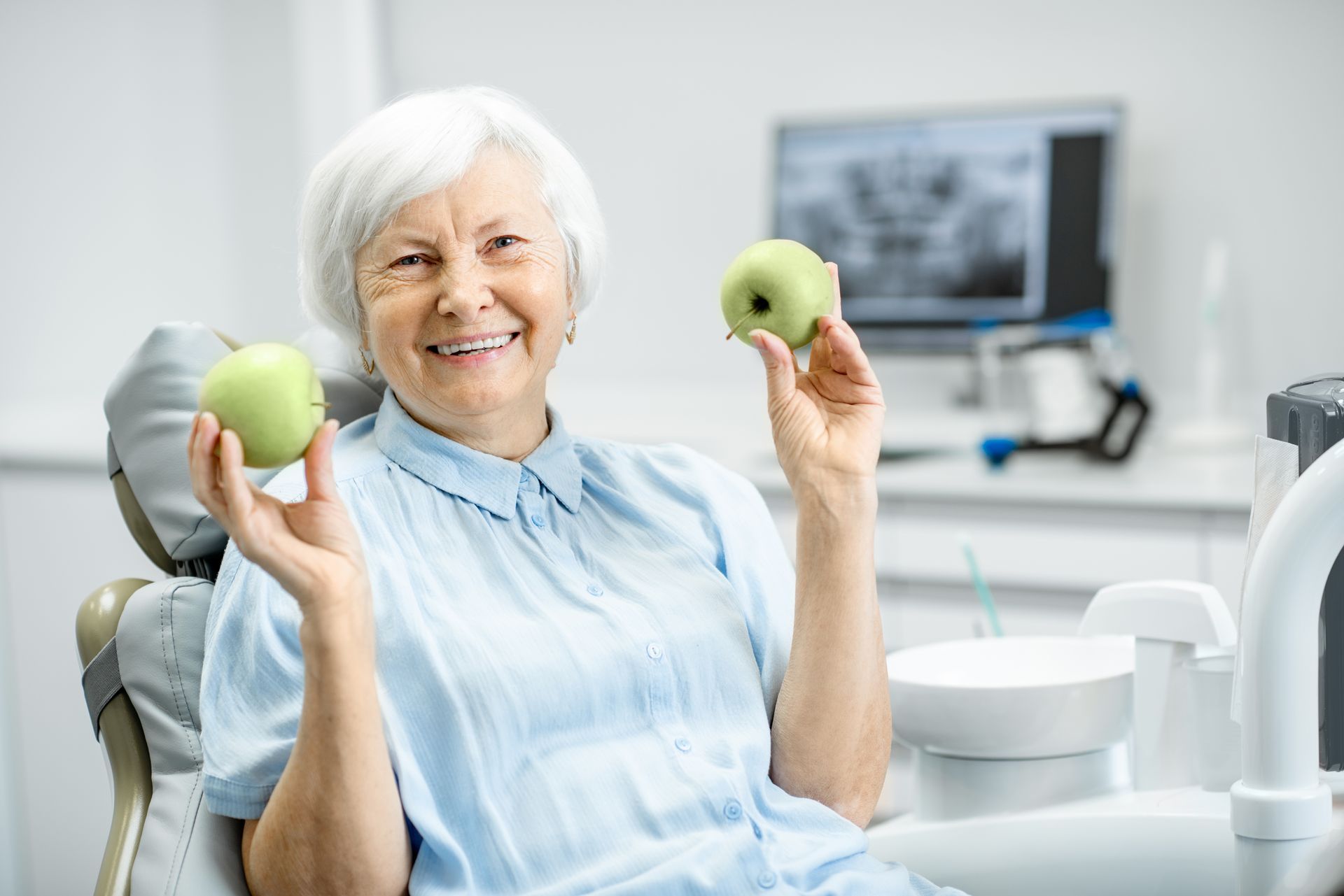 A person sitting in a dental chair, smiling and holding a green apple in each hand, with dental X-rays on a monitor.