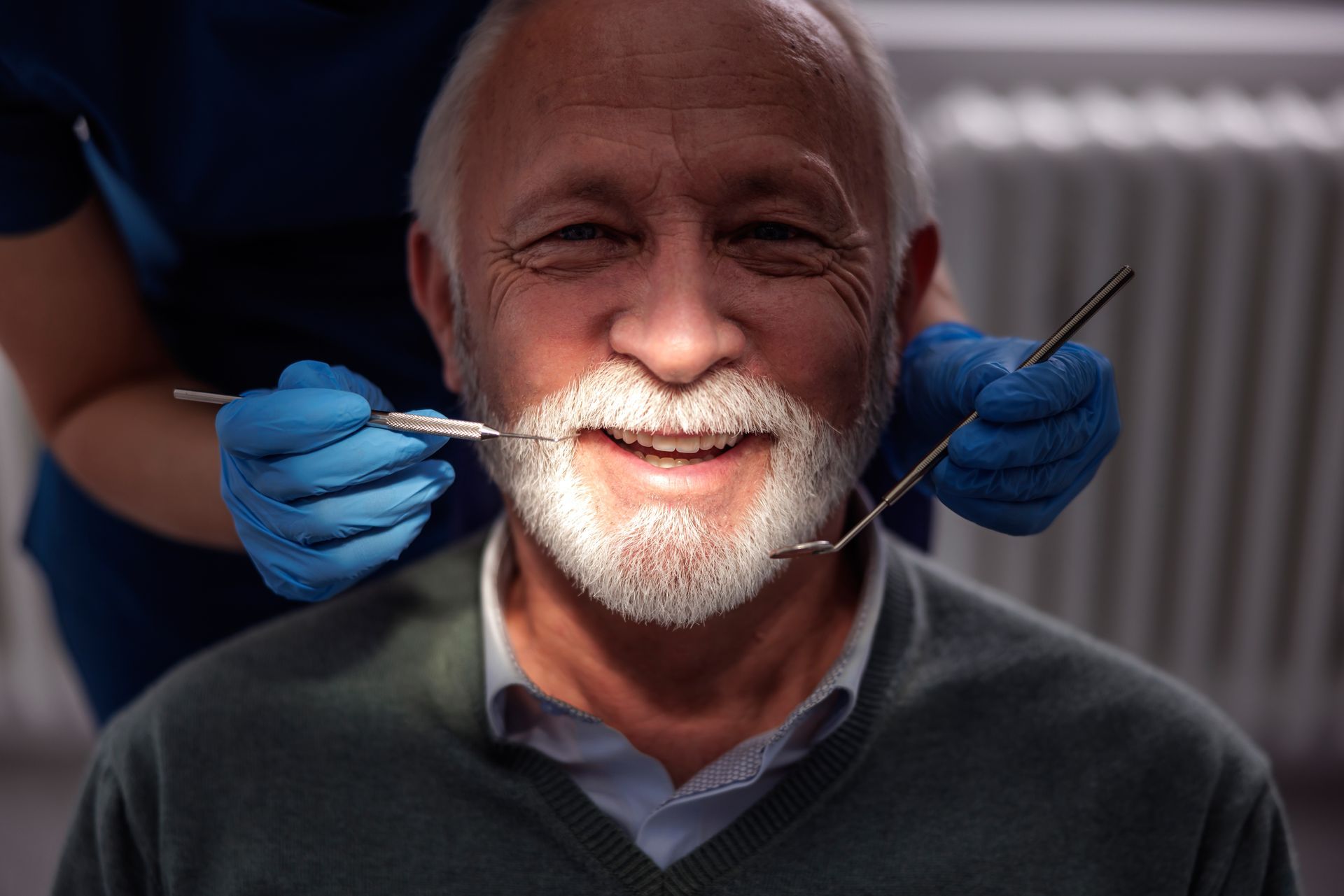 A dentist wearing blue gloves uses dental tools to examine a smiling patient's teeth in a clinical setting.
