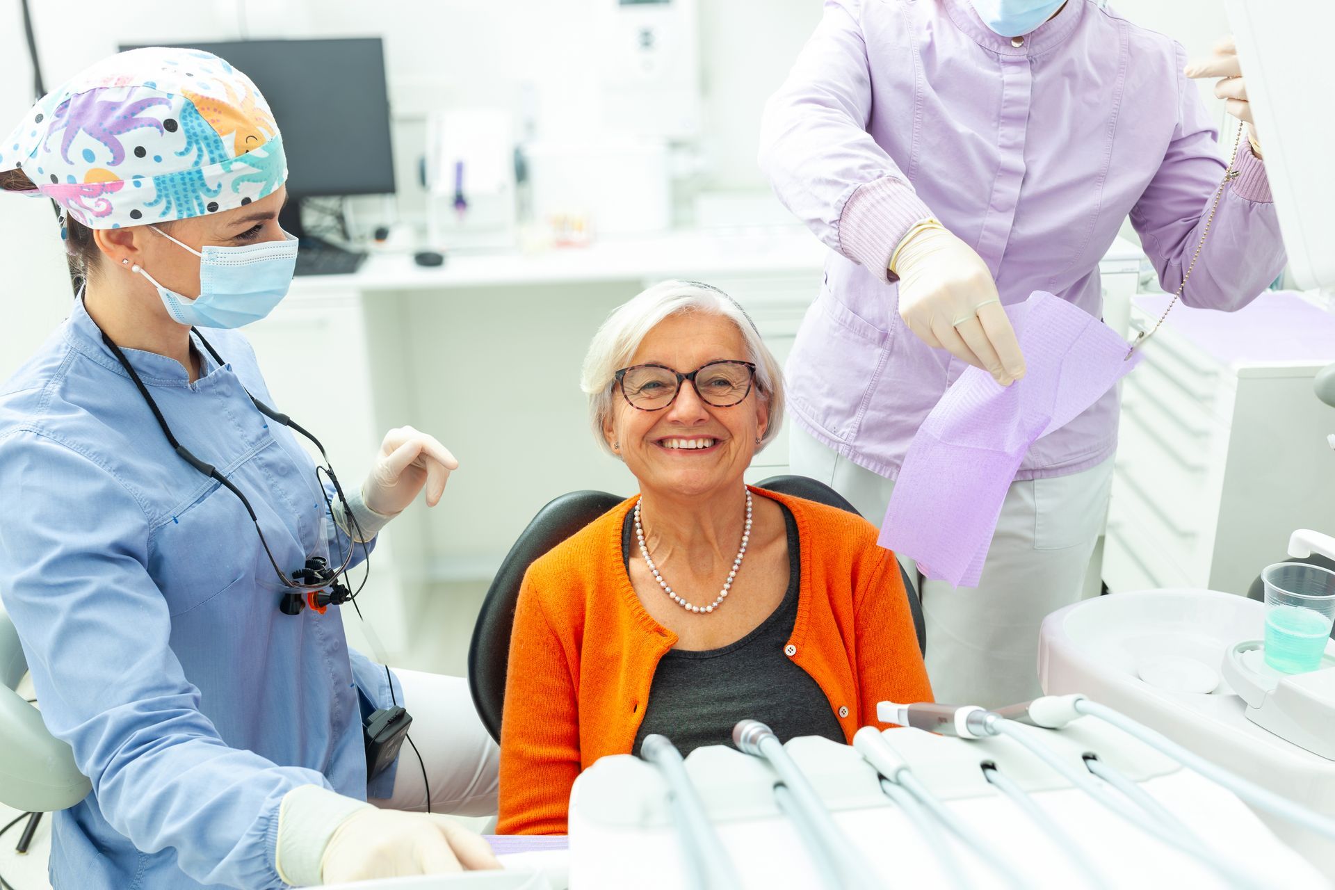 Woman smiles at dentist. Dental assistant prepares bib. Dental office setting.