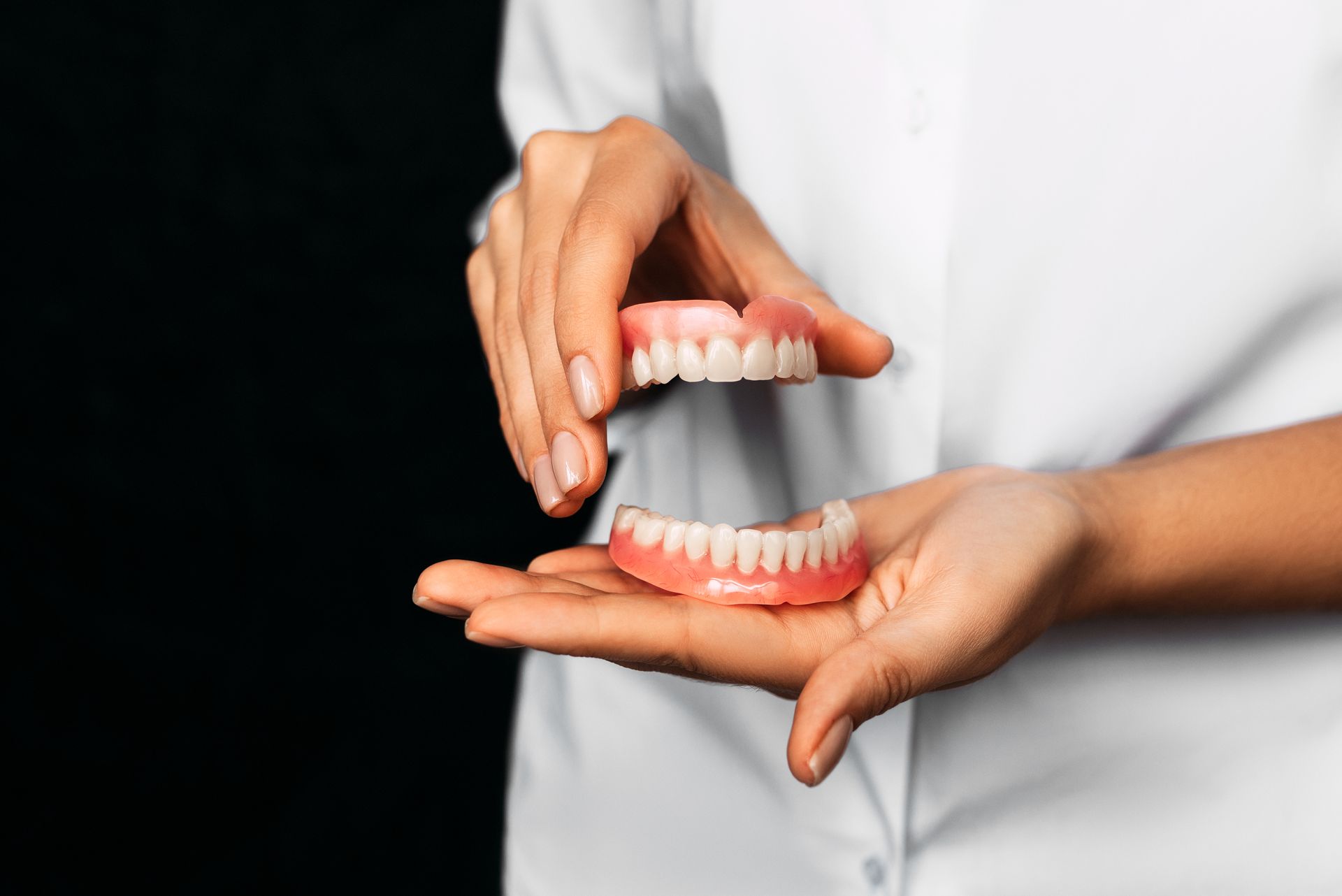 Person holding upper and lower dentures, pink gums, white teeth, against a dark background.