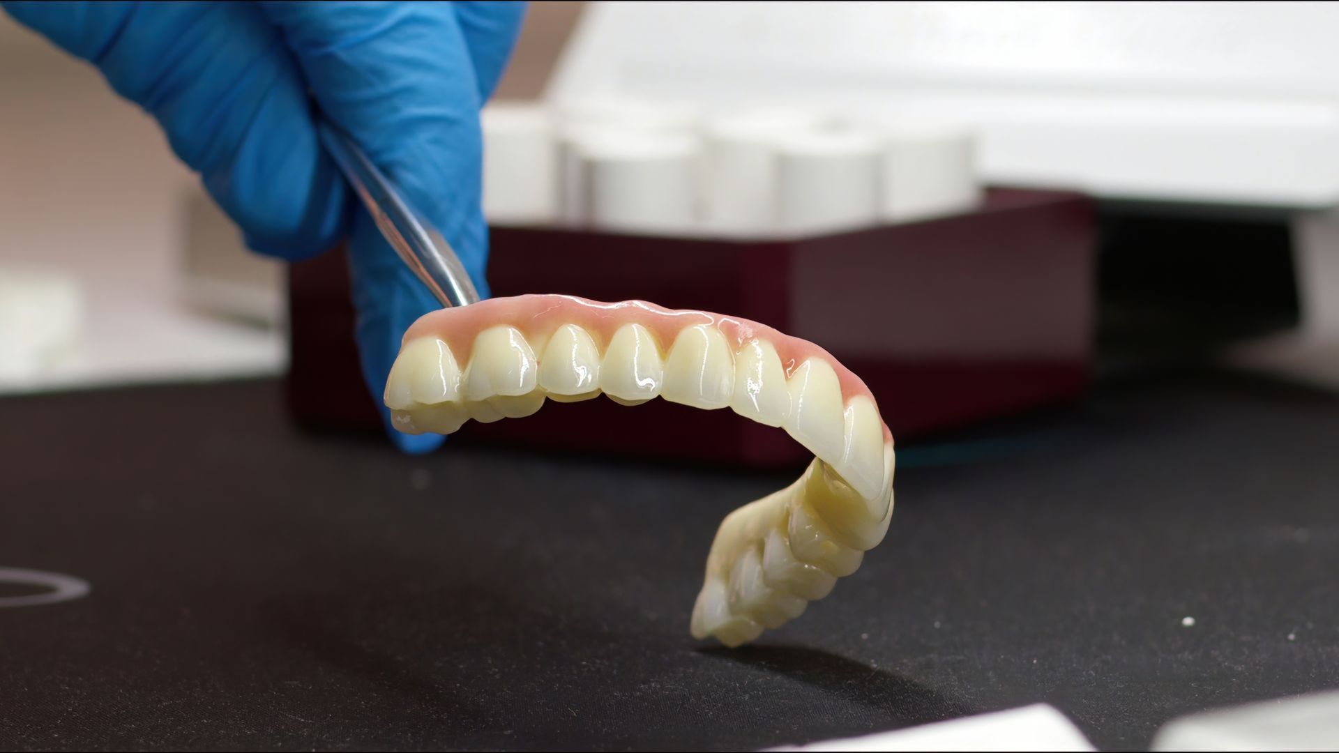 Dentures held by tweezers, showing upper teeth with pink gum and white teeth.