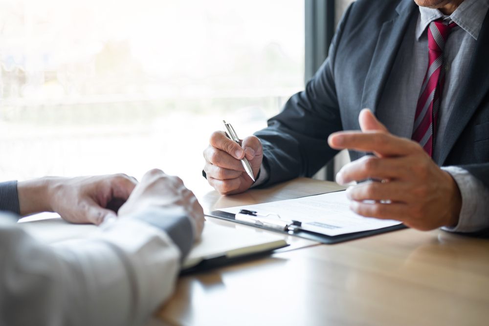 Two Men in Suits at a Table, One Gesturing With Pen Over Paperwork, Discussing — People Assured Partners in Cannonvale, QLD
