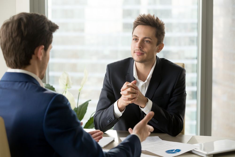 Two Men in Suits at a Table, One Gesturing and Speaking the Other Listening — People Assured Partners in Mackay, QLD