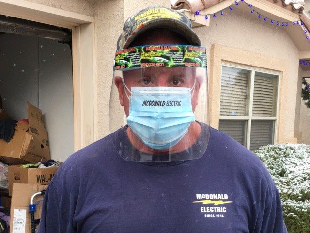 Man in blue shirt, face mask, and face shield stands outside a house with boxes; snowy ground.