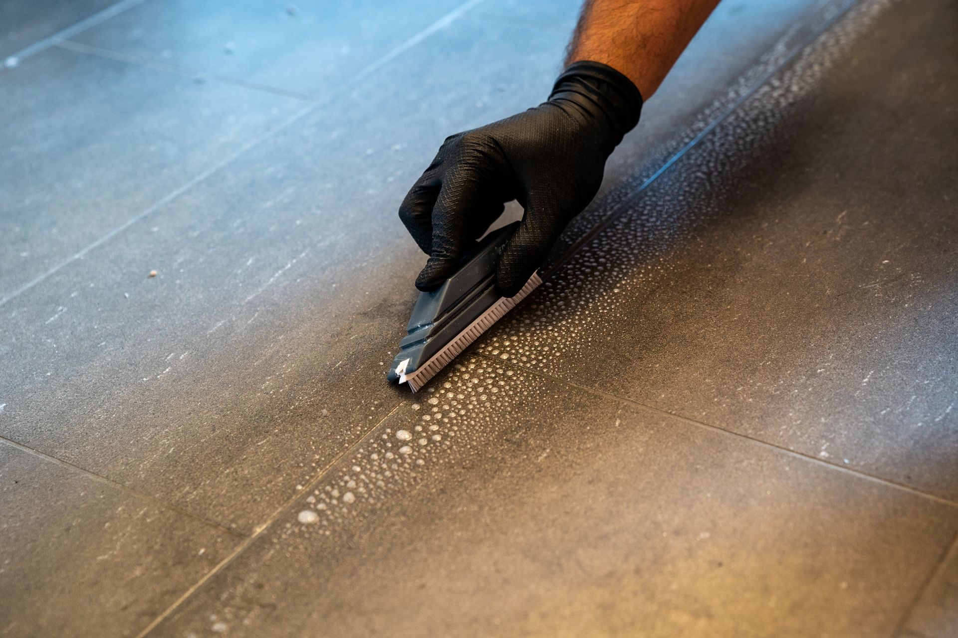 A person wearing black gloves is cleaning a tile floor with a sponge.