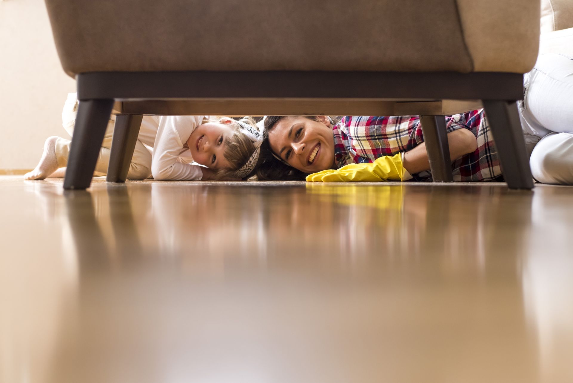 A woman and a child are cleaning the floor under a couch.