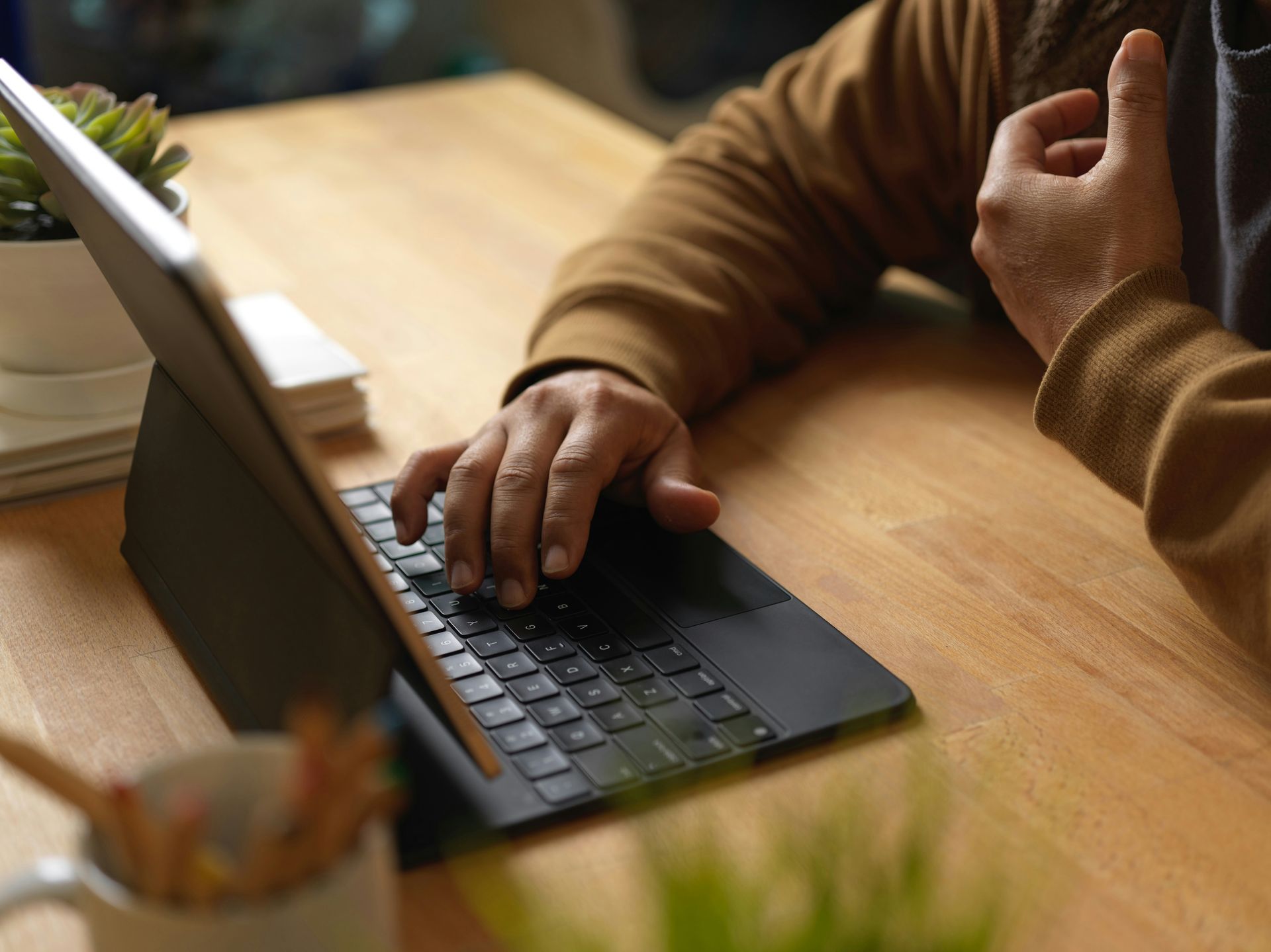 Person typing on a black tablet with attached keyboard, seated at a wooden desk, near a small plant.