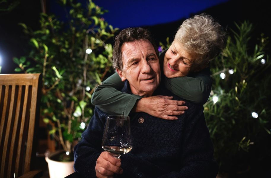 Older couple embracing outdoors at night; woman hugging man from behind, holding wine glass.