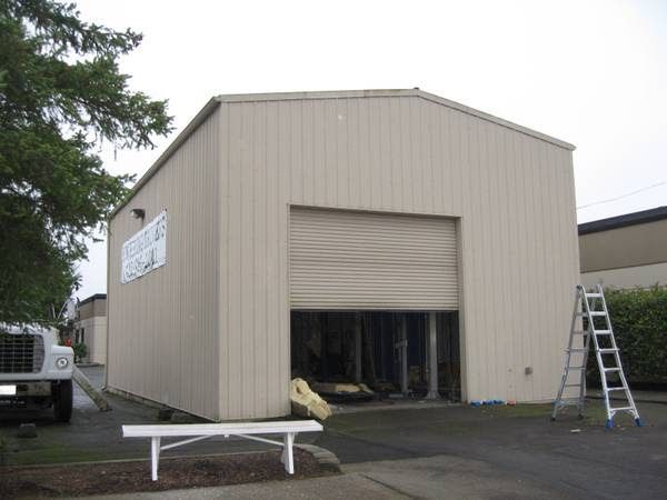 Tan metal building with an open garage door and a white bench. A ladder leans against the side.