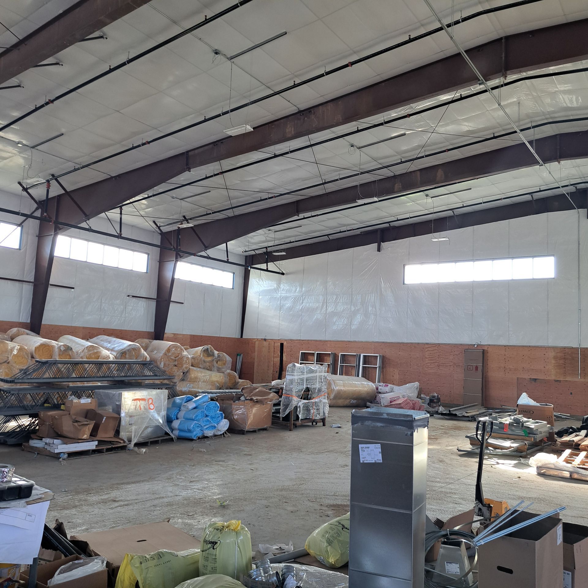 Large, open warehouse interior with metal beams, white walls, scattered pallets of supplies, and a water fountain.