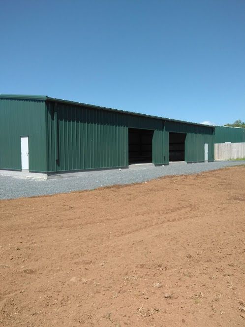 A long, green industrial storage building with two open bays, set on a gravel base next to a dirt lot under a blue sky.