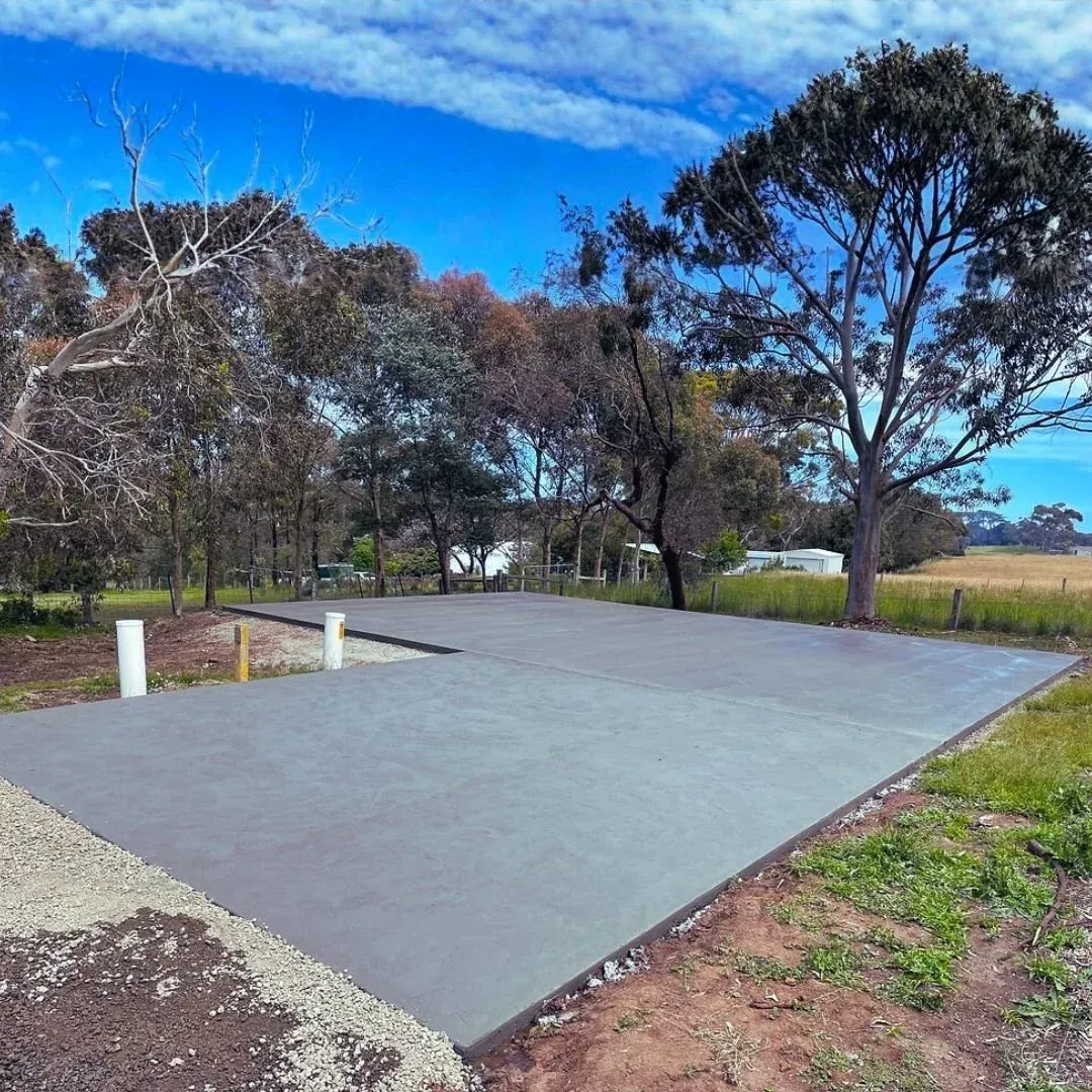 A freshly poured concrete slab foundation sits in a rural field near trees under a bright blue sky.