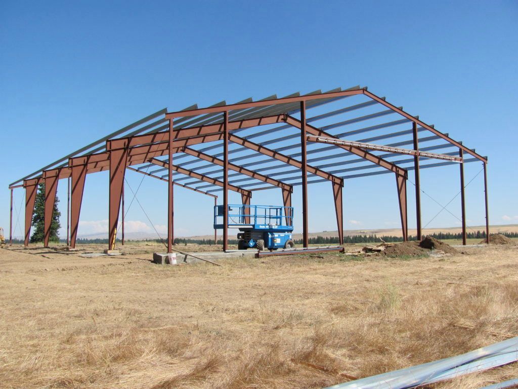 A metal building frame under construction in a dry, open field with a blue lift parked inside under a clear blue sky.