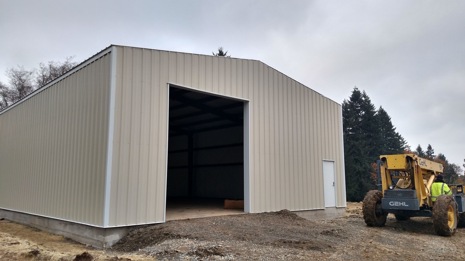 Tan metal building with large open doorway and a small door, with a yellow construction vehicle.