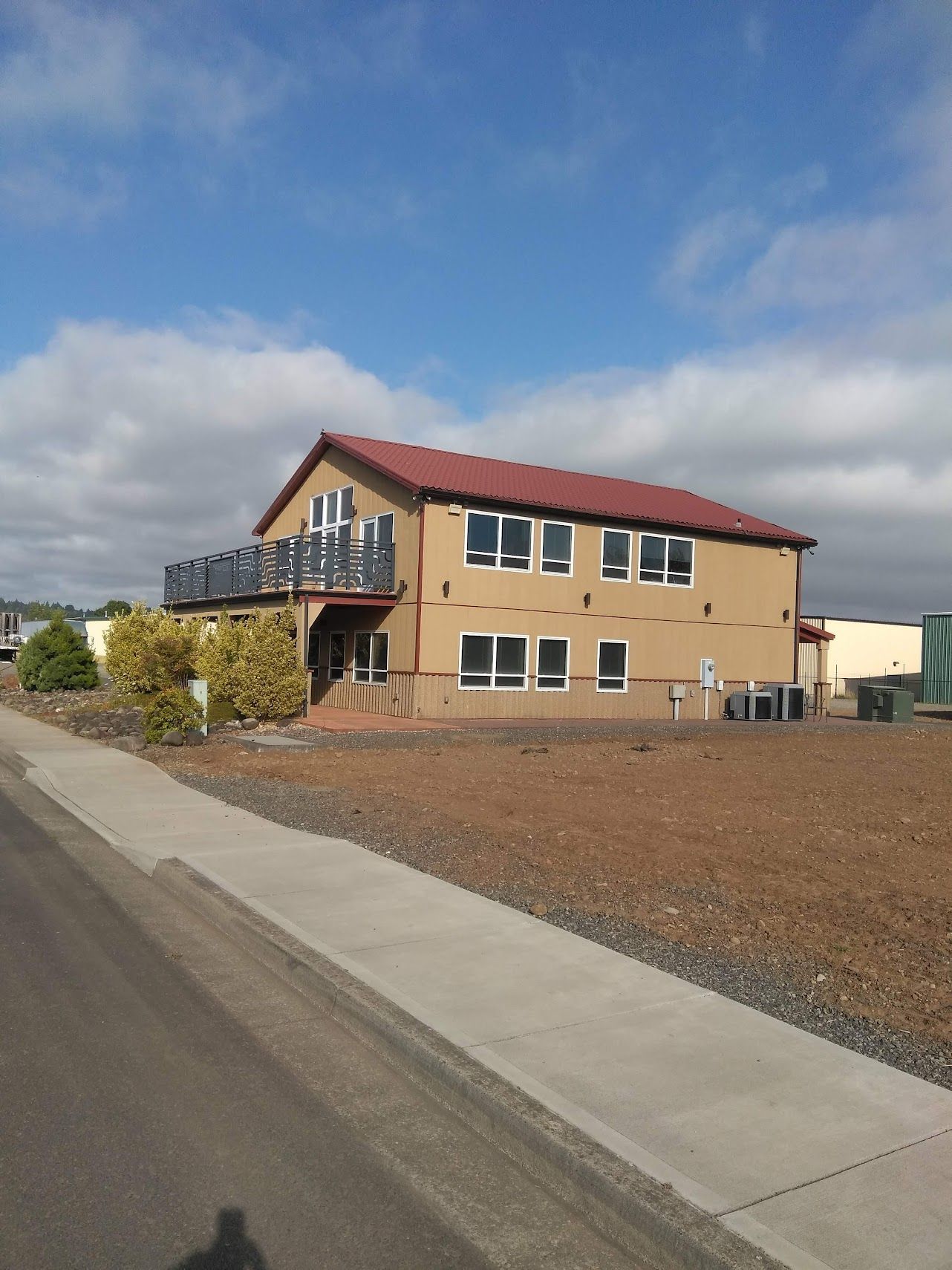 A two-story tan building with a red gabled roof and a second-story balcony sits next to a sidewalk on a sunny day.