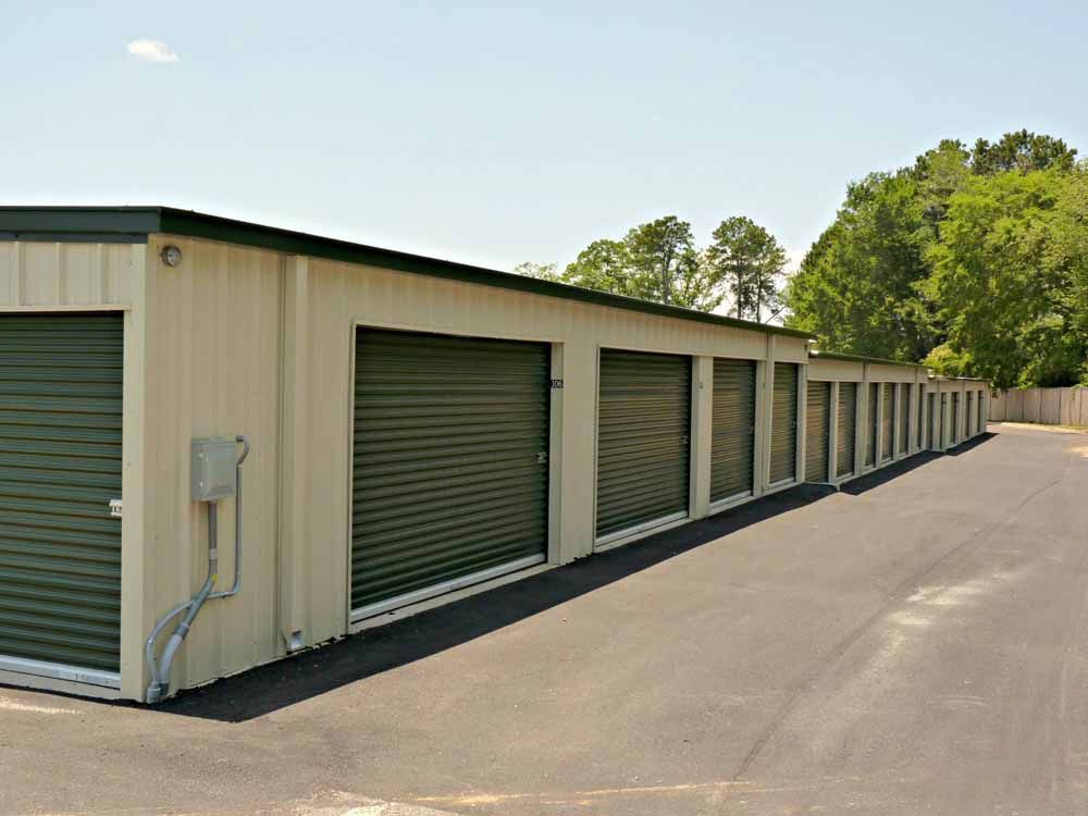 Storage units with green doors along a paved drive under a clear sky.