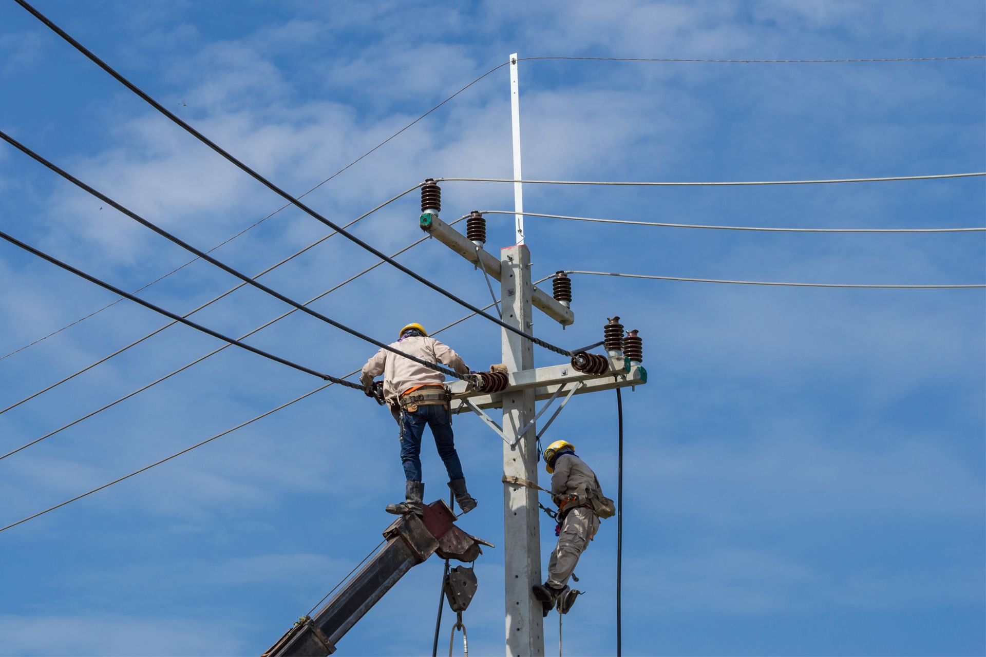 Two utility workers on a power pole repairing electrical lines against a blue sky.