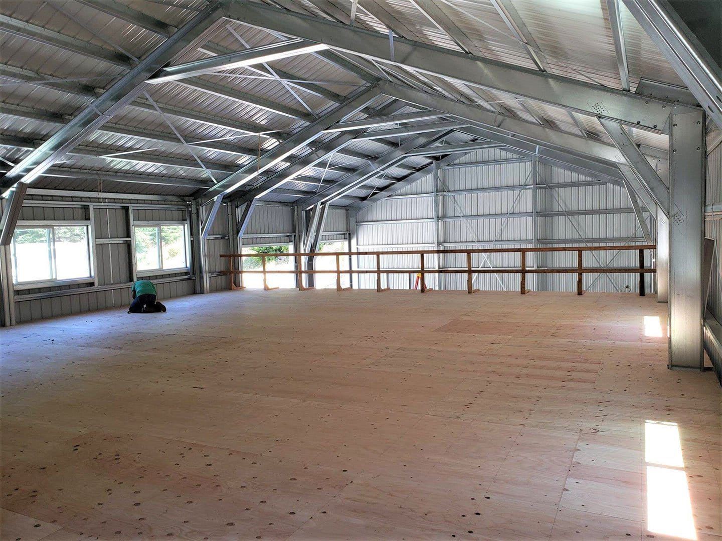 An interior view of a wide, unfinished metal shed loft with a plywood floor, exposed steel rafters, and wood railings.