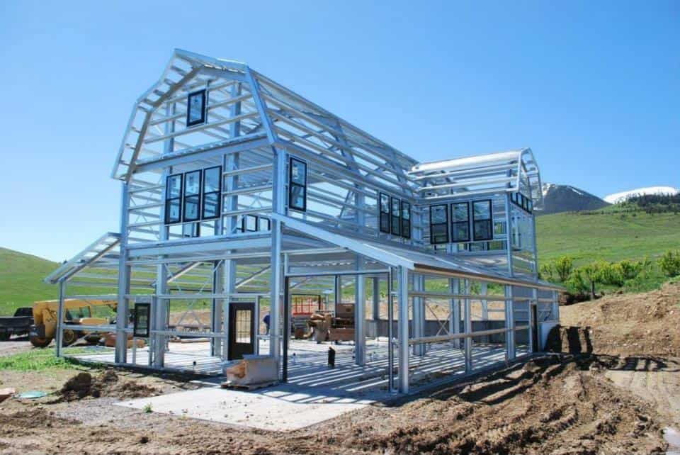 Steel frame of a two-story barn-style house under construction, with windows and a clear blue sky.