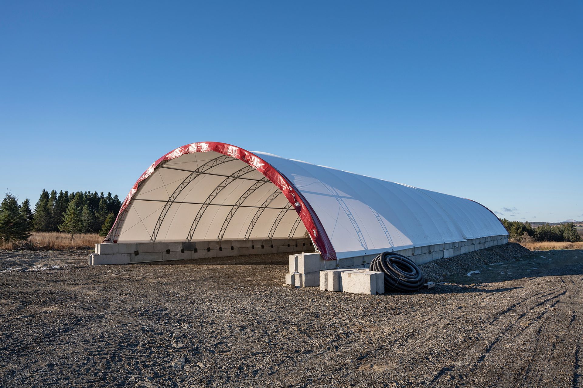 White arched-roof structure with red trim, set on concrete blocks in a gravel field under a blue sky.