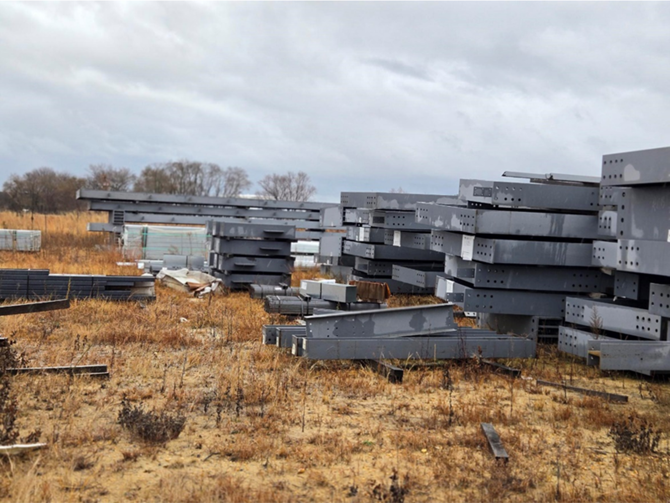 Stacked steel I-beams lay in a field under a cloudy sky.