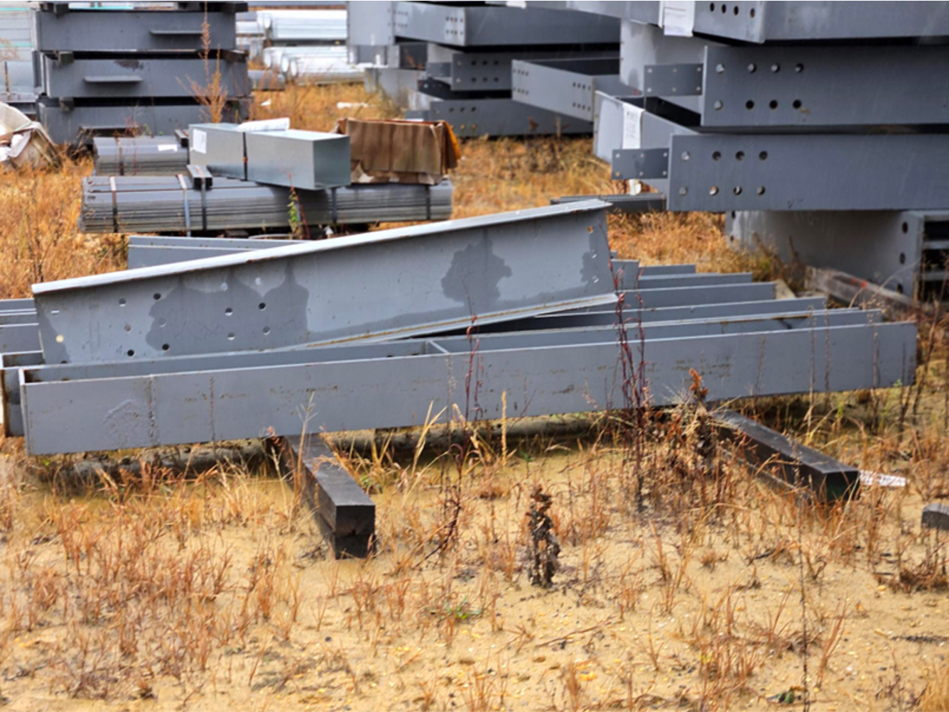 Metal beams and supports lying on dry, brown ground at a construction site.
