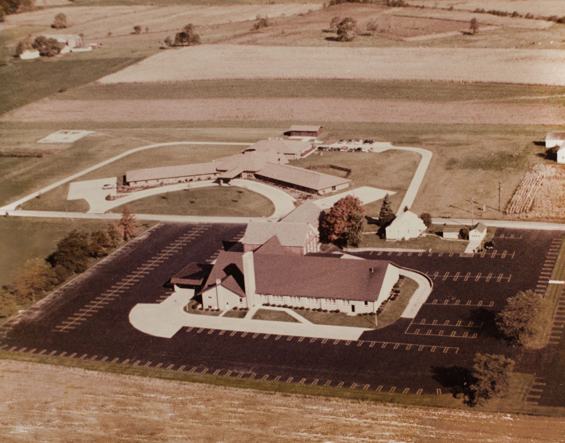 Aerial view of a church and surrounding buildings with a large parking lot, set in a rural landscape.