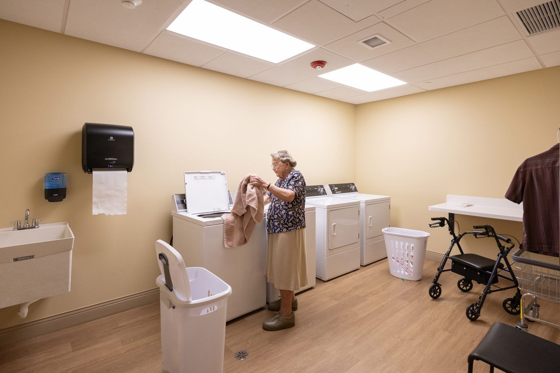 Elderly person doing laundry in a laundry room, with a washing machine, sink, and walker.