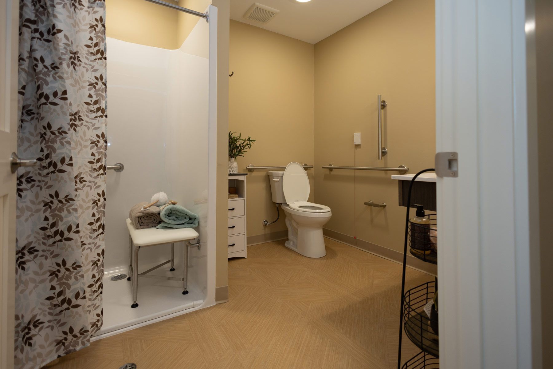 Accessible bathroom with shower, toilet, and grab bars; beige walls, neutral floor, shower chair, and shower curtain.