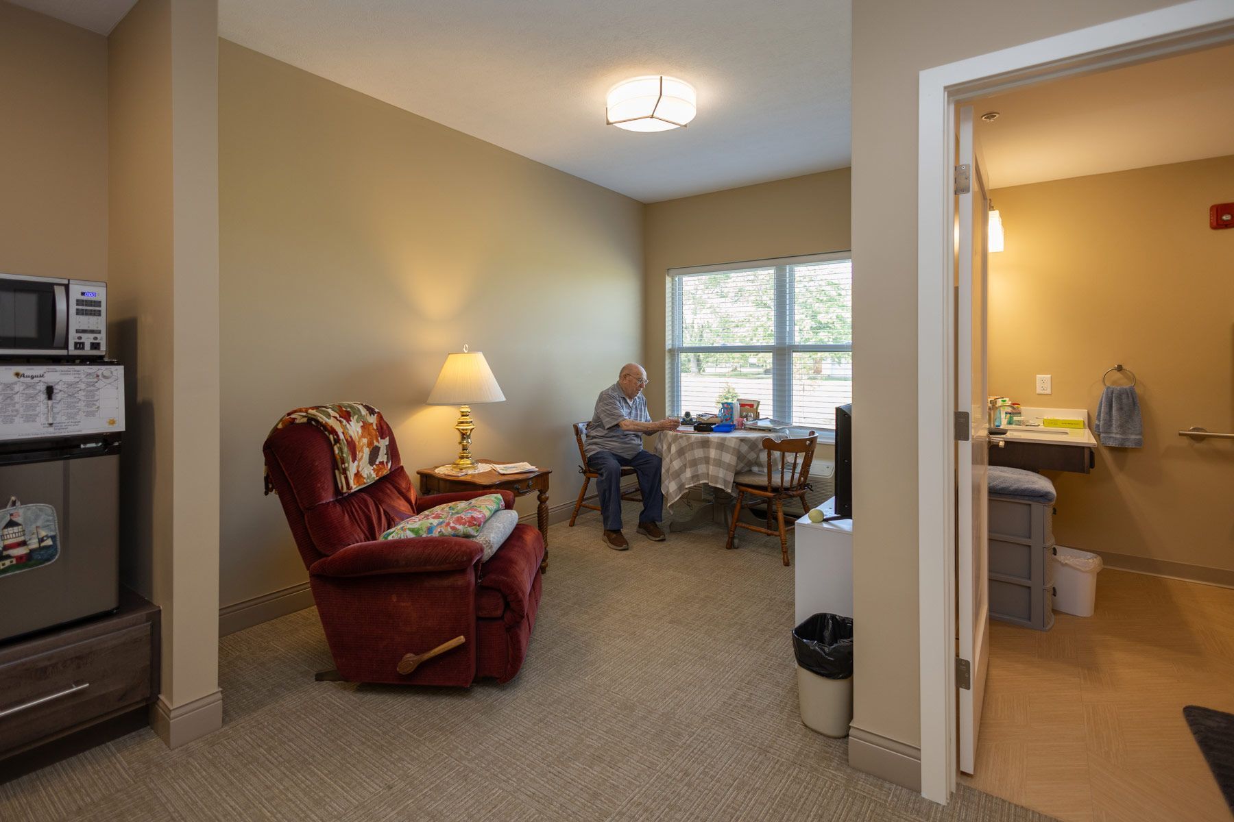Senior in a room with a recliner, table, and microwave. Bathroom visible in the doorway.