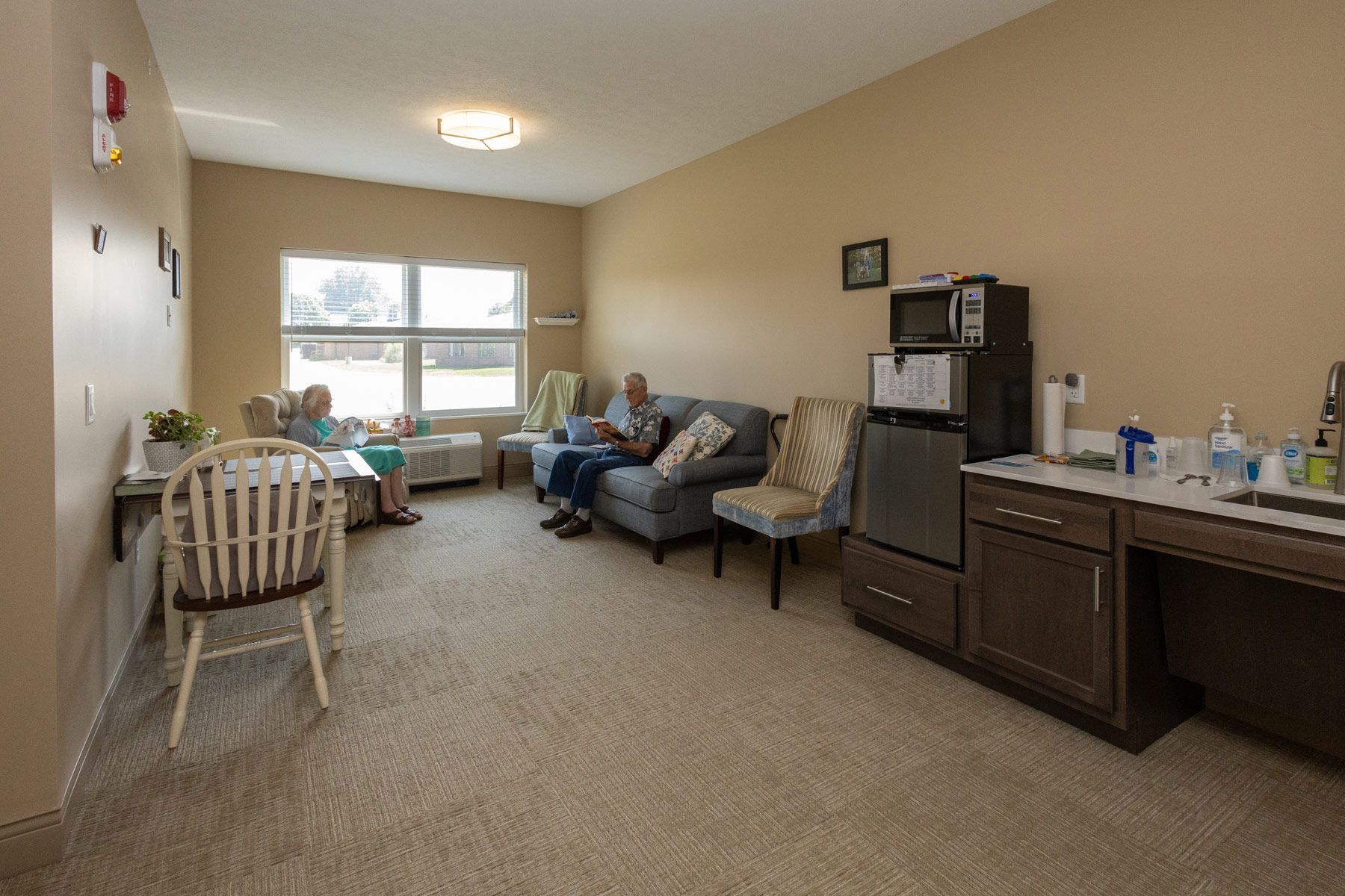 Interior of a living area in a care facility with two people; one is seated at a table, and another on a couch.