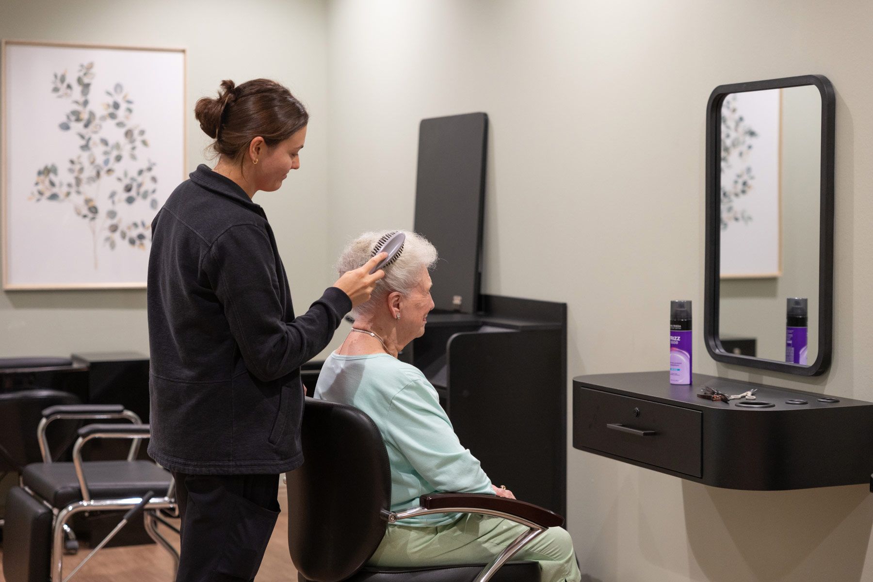 Hairdresser brushing a seated person's hair in a salon.  Black counter, mirror, and equipment, light green walls.