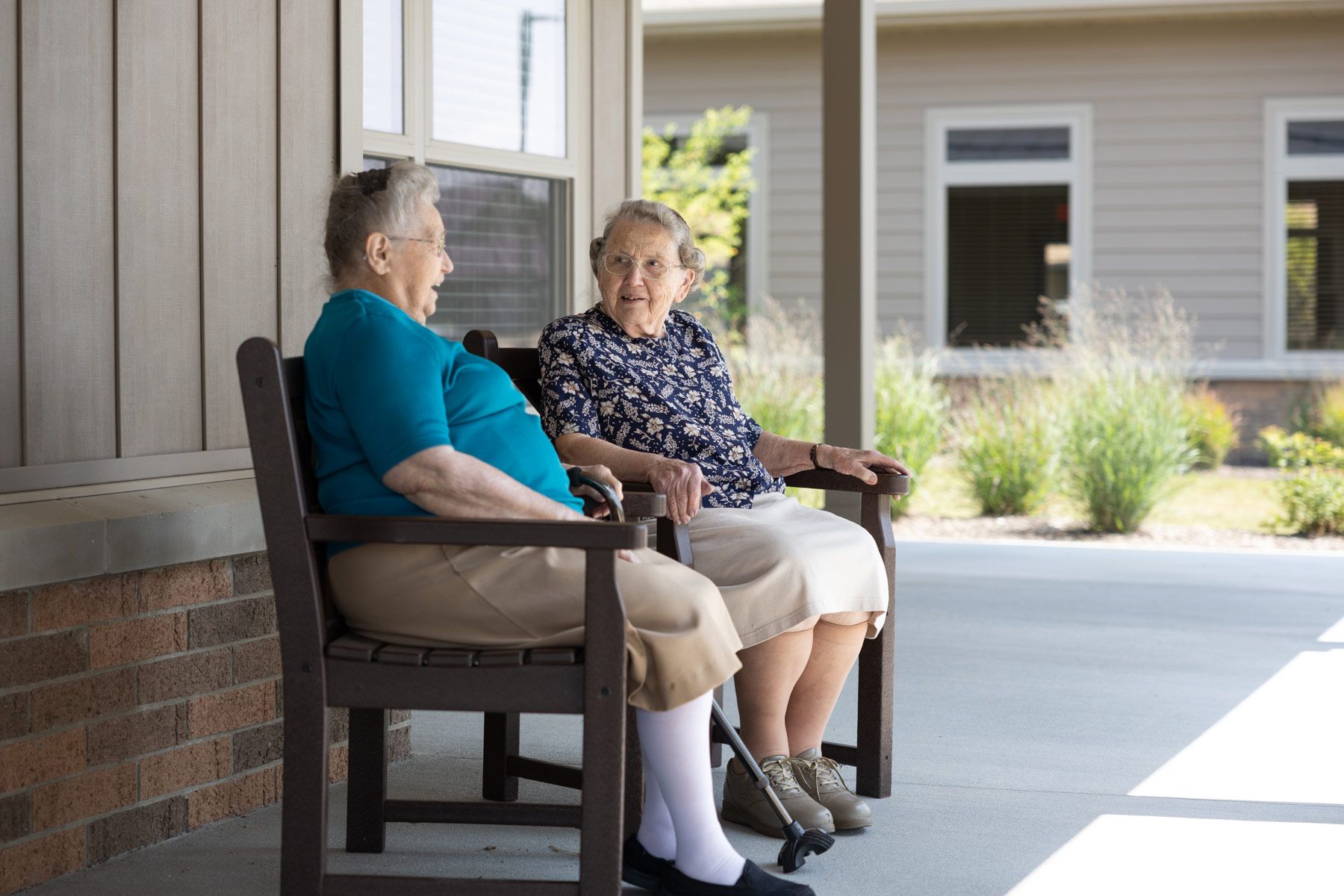 An older person and a person sit on chairs on a porch of building number 38. The weather appears sunny.