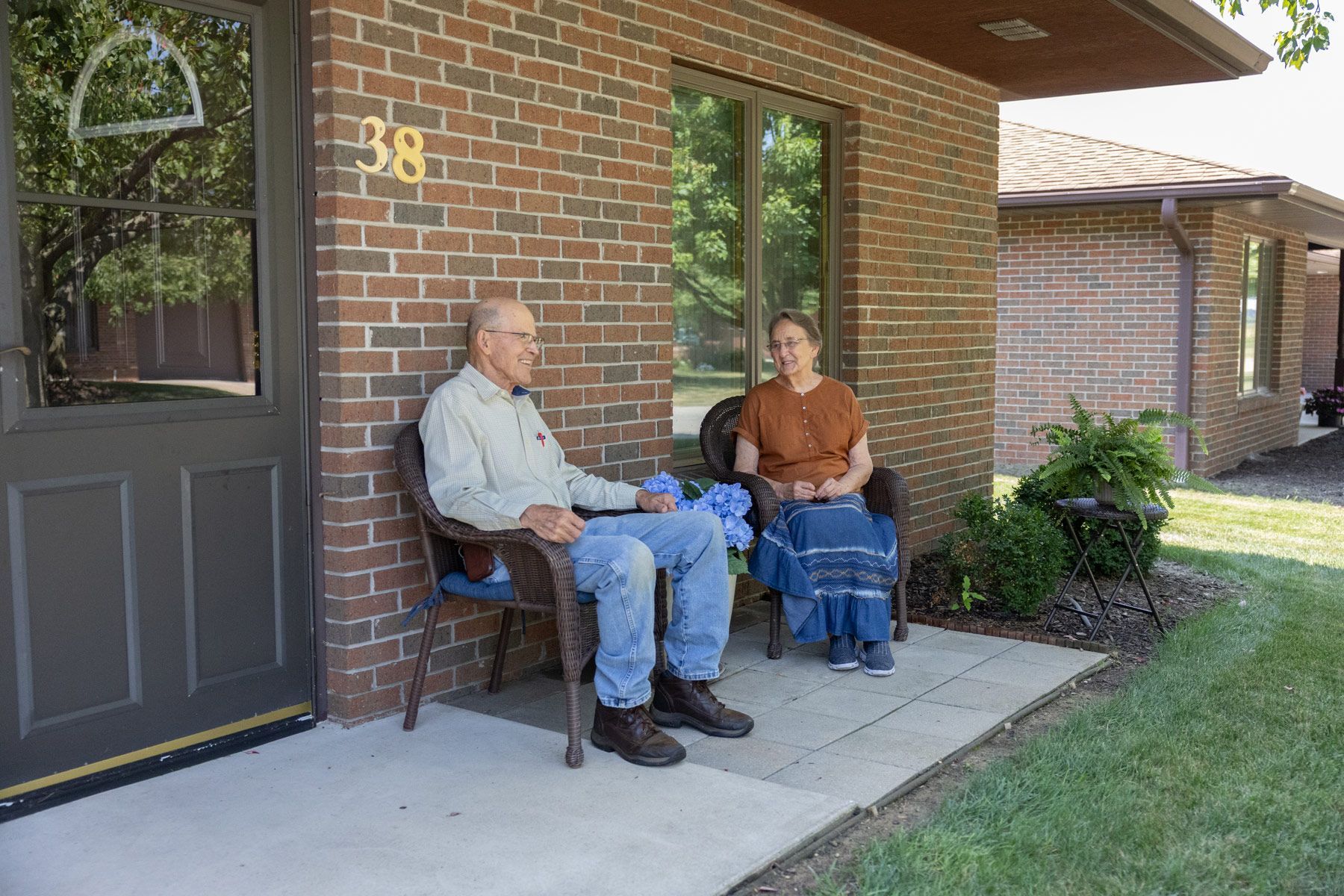 An older couple sitting on chairs on a porch outside a brick building, smiling, sunny day.