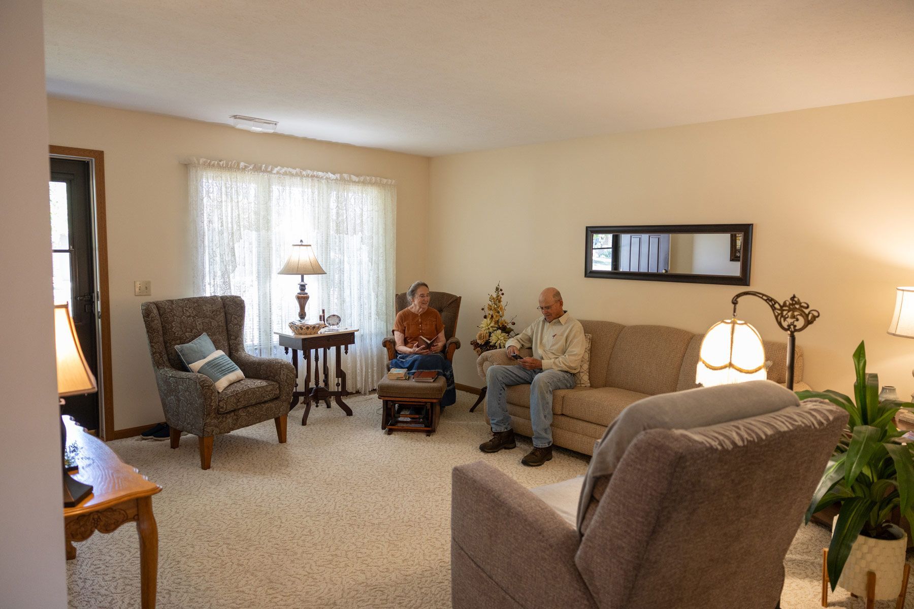 Two people sit in a living room with tan walls, a couch, and chairs.