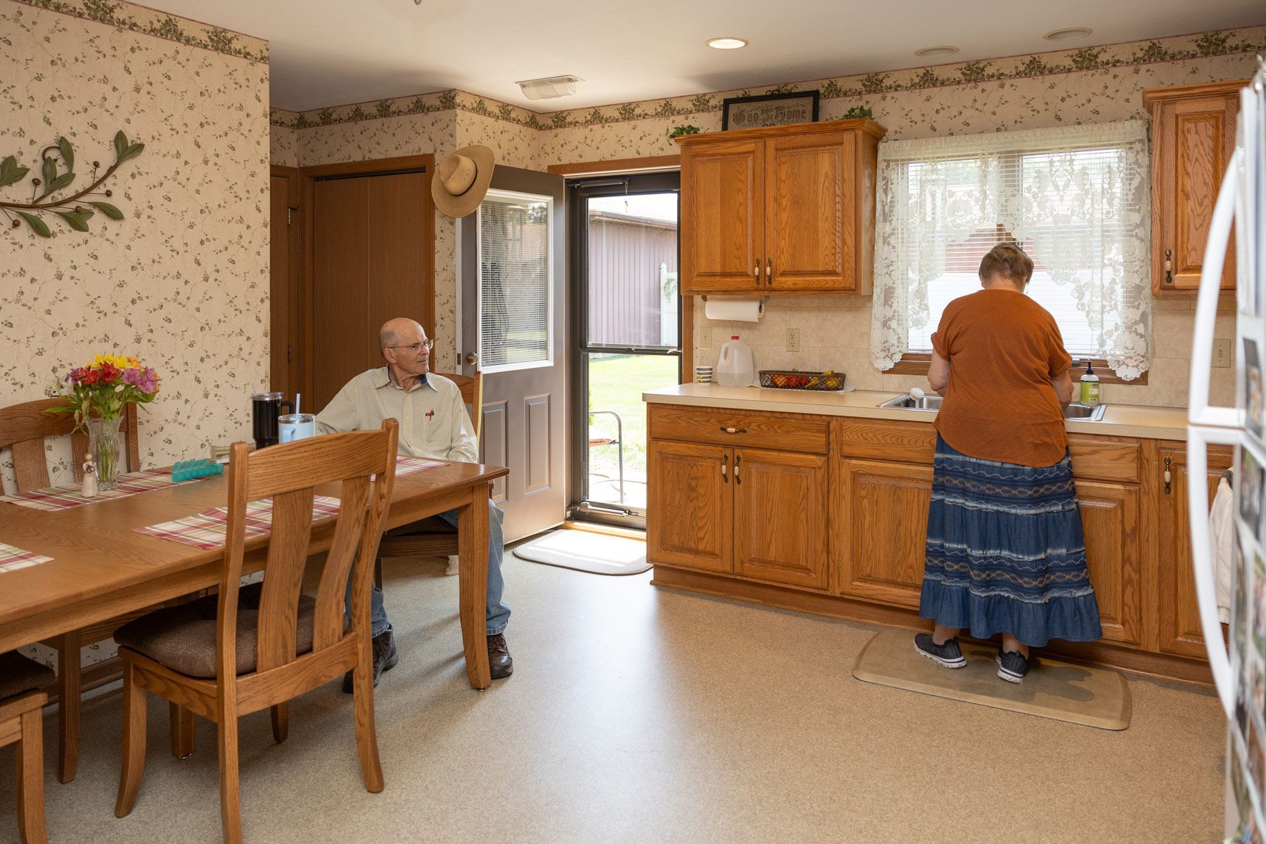 Kitchen scene with a person washing dishes and another seated at a table.