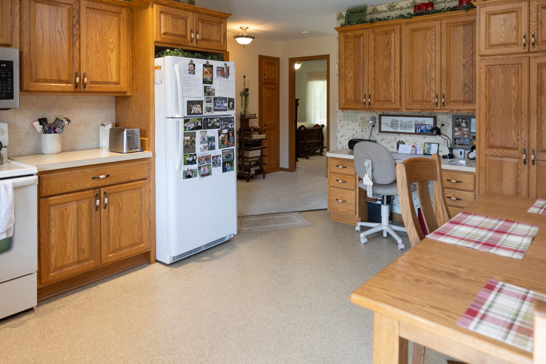 Kitchen with wooden cabinets, refrigerator, and a table. A desk and a doorway are visible.
