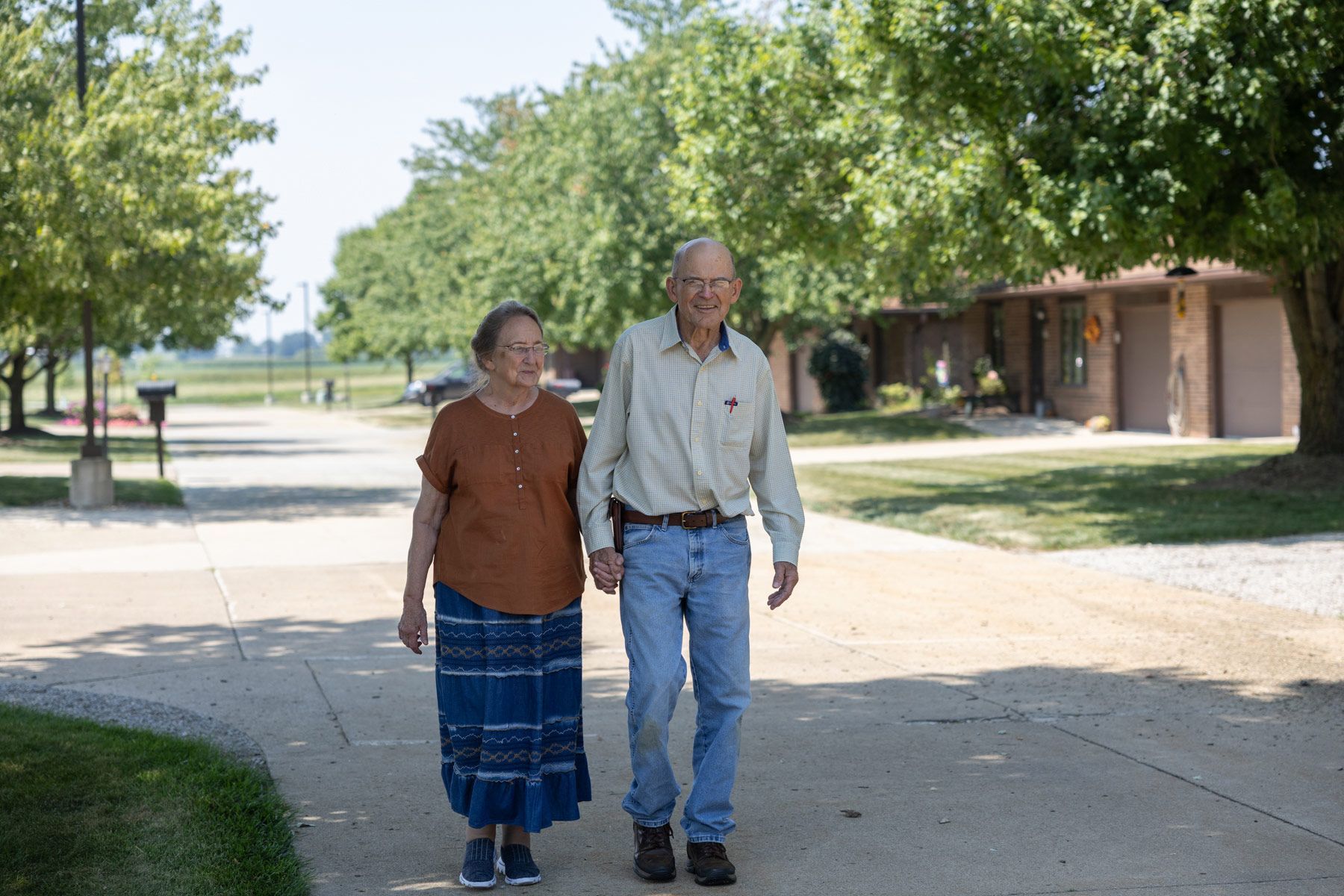 Elderly couple walks hand-in-hand on a paved path outdoors, under the sun.