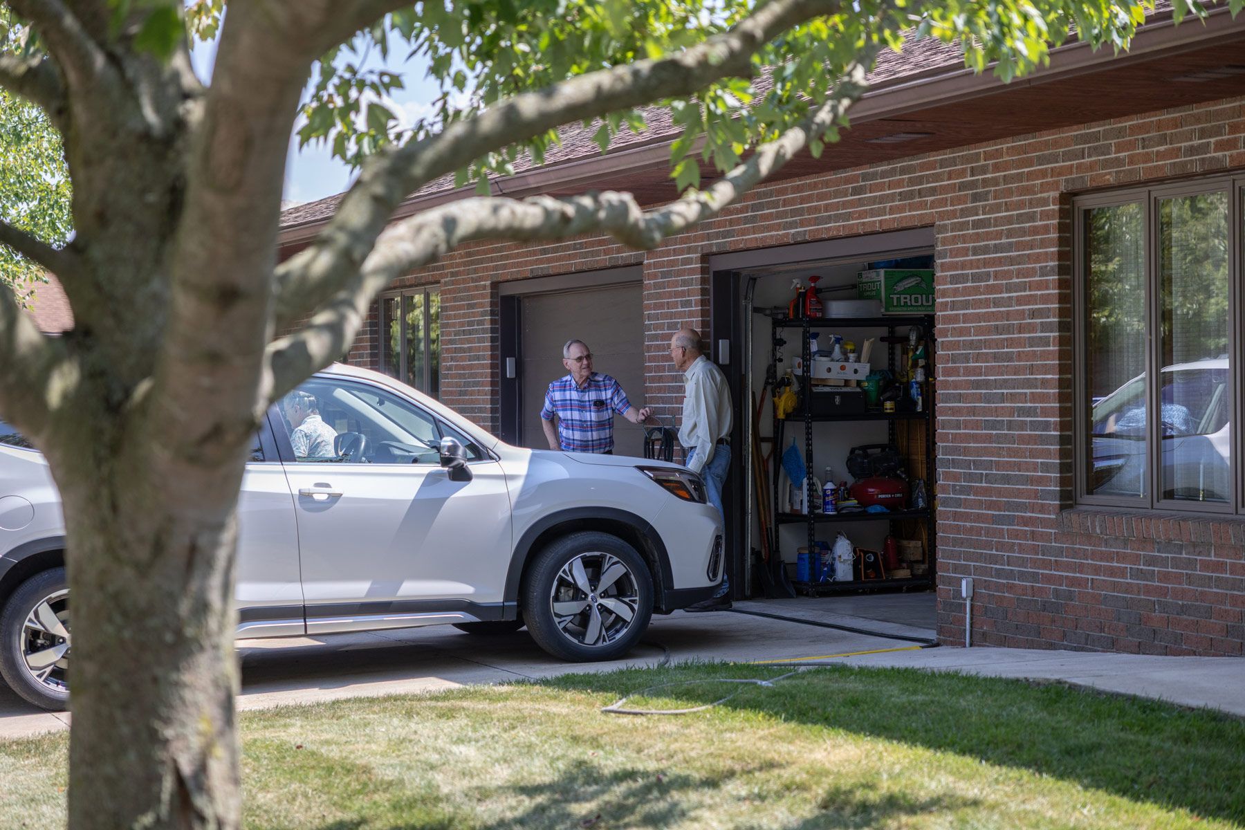 White SUV parked near a brick garage. Two men stand inside the open garage door, facing each other. Tree in foreground.
