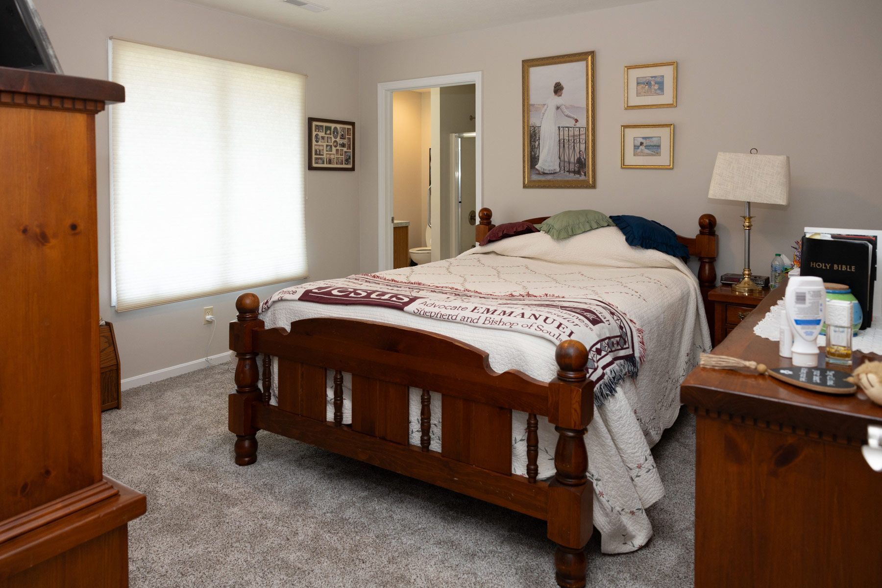 Bedroom with a wooden bed, dresser, and a window with blinds. The room has neutral-colored walls and carpet.