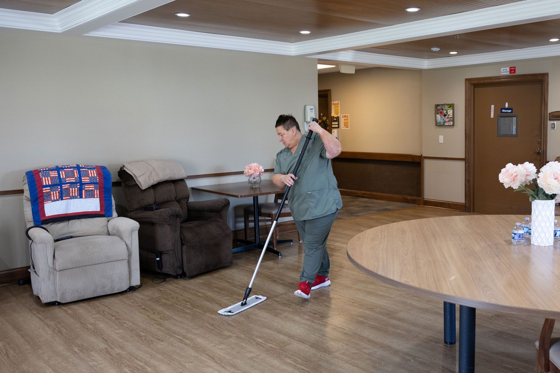 Person mopping a light-colored floor in a room with a recliner, tables, and a vase of flowers.