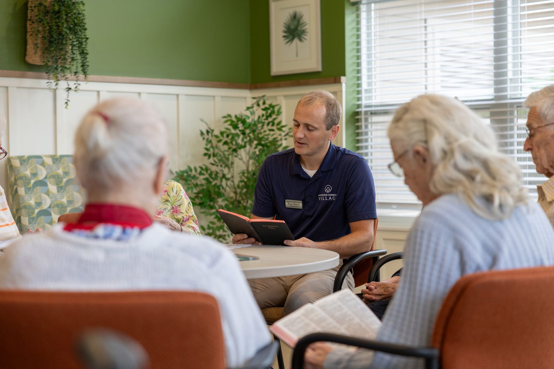 Group of people in chairs around a table, one person is reading a book. Interior setting, well lit.