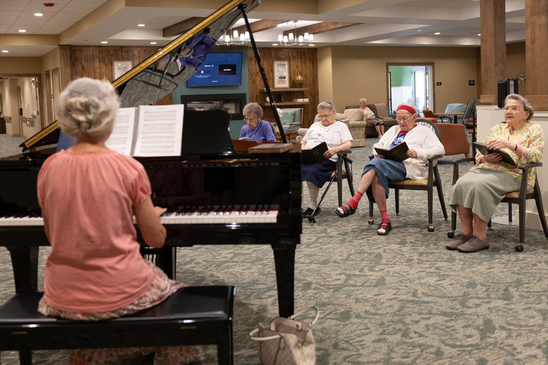 Woman playing piano, four others singing, in a room with chairs and music sheets.